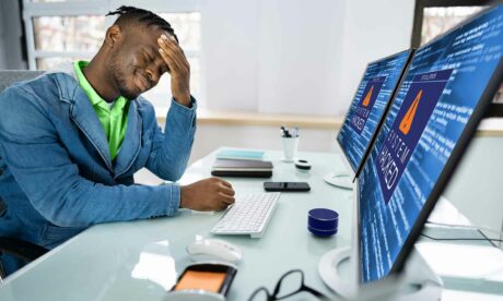 The feature image for an article on the negative effects of cyber attacks on businesses and individuals (consumers). It's a photo of a grimacing man holding his head with the words "System Hacked" showing on his computer monitors.